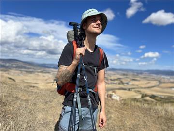 Ein Mann in Italien auf dem Jakobsweg. Blauer Himmel und die Landschaft sind unscharf im Hintergrund zu sehen. Er trägt einen Anglerhut und und zwei Wanderstöcke in der rechten Hand. Er lächelt.  TV-Aussteiger Tobi Schlegl spricht im Interview über den mehr als 700 Kilometer langen Jakobsweg, den er mit seiner Mutter gegangen ist, welche Rolle Hape Kerkeling dabei spielte und sein Leben als Notfallsanitäter.
