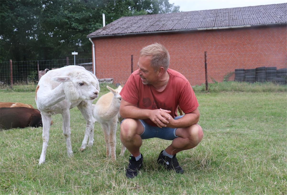Ein Mann hockt auf einer Weide, hinter ihm zwei Alpakas.