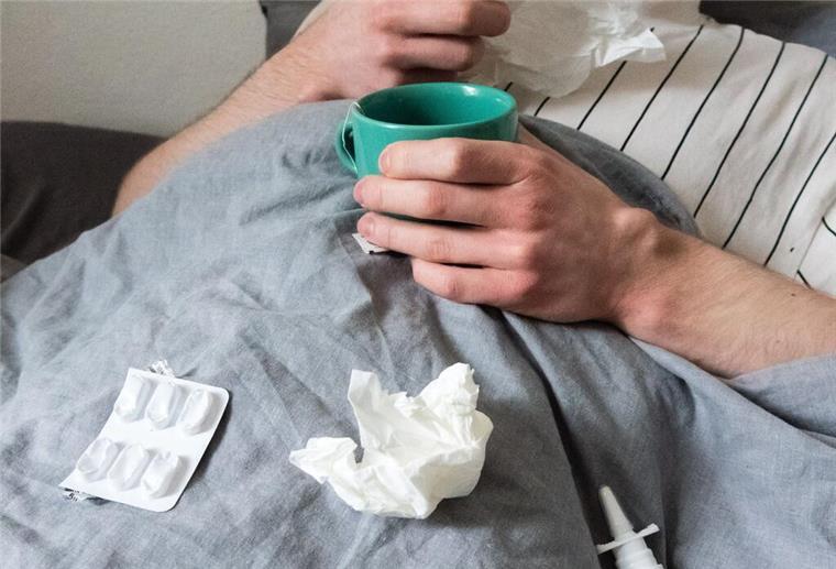  Ein Mann liegt mit Taschentüchern, Teetasse, Nasenspray und Tabletten im Bett.