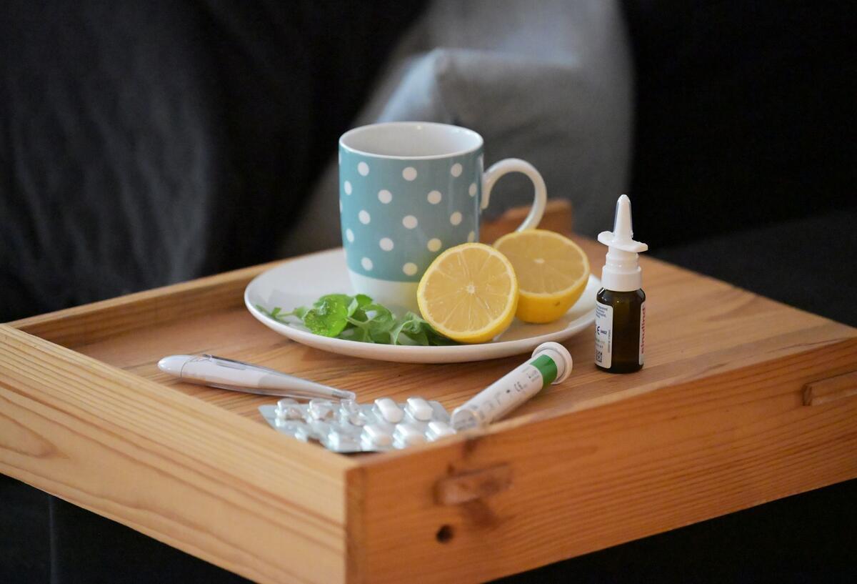Anstieg bei Corona und Erkältungen: So ist die aktuelle Lage Eine Tasse mit dampfendem Tee samt Zitronenhälften steht auf einem Tablett, daneben befinden sich Nasenspray, Fieberthermometer und Medikamente. (gestellte Szene)