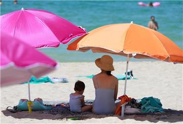 Eine Frau sitzt neben ihrem Kind unter einem Sonnenschirm an einem warmen Sommertag am Strand. 