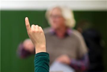 Ein Kind meldet sich, im Hintergund steht ein Lehrer vor einer grünen Tafel.